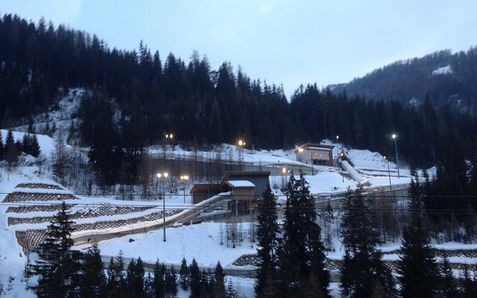A snow-covered bobsled track winds through a forested mountainside at dusk, lit by lamps