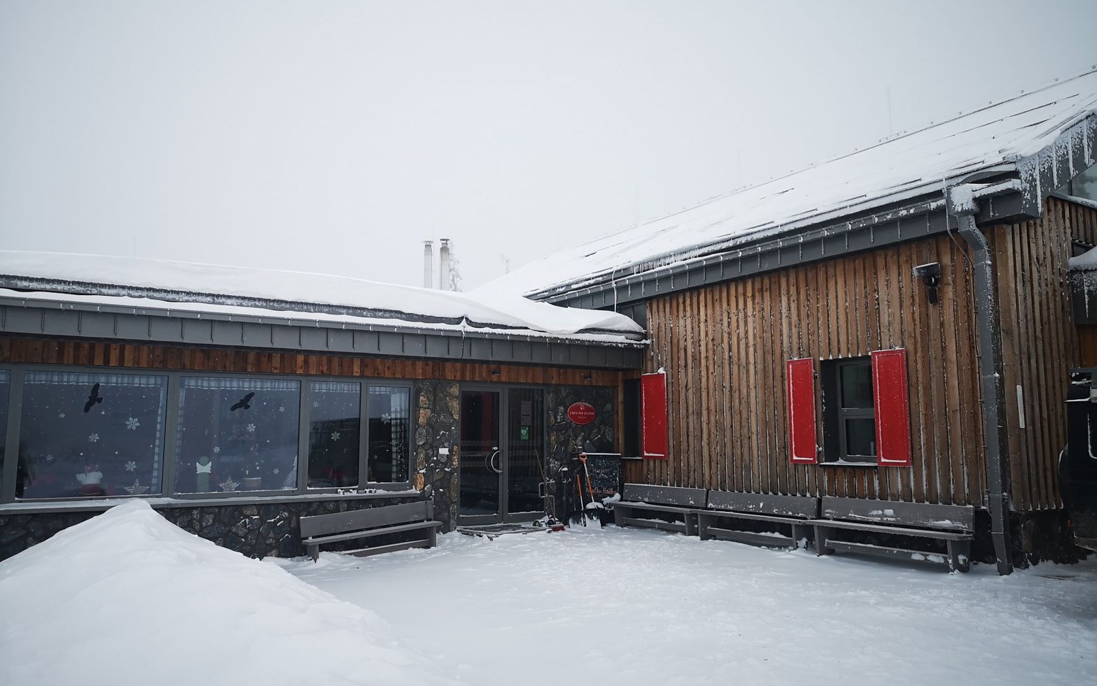 Snow-covered wooden mountain lodge with red window shutters and benches out front