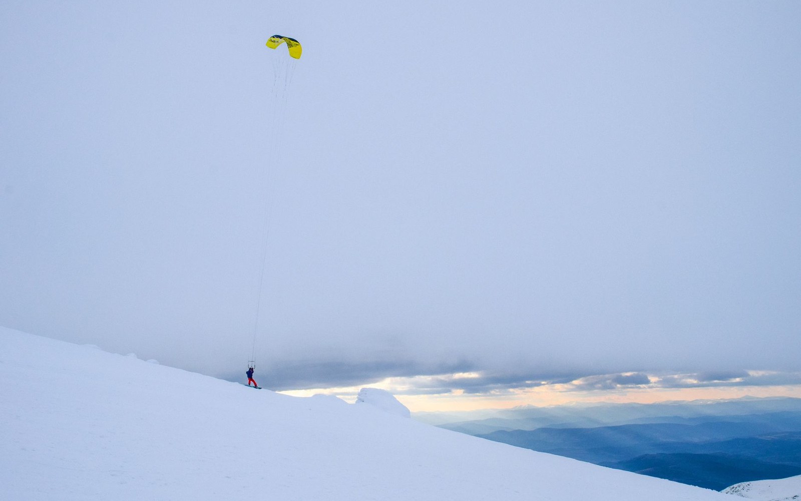A skier tethered to a yellow kite descends a snowy mountain slope at dusk