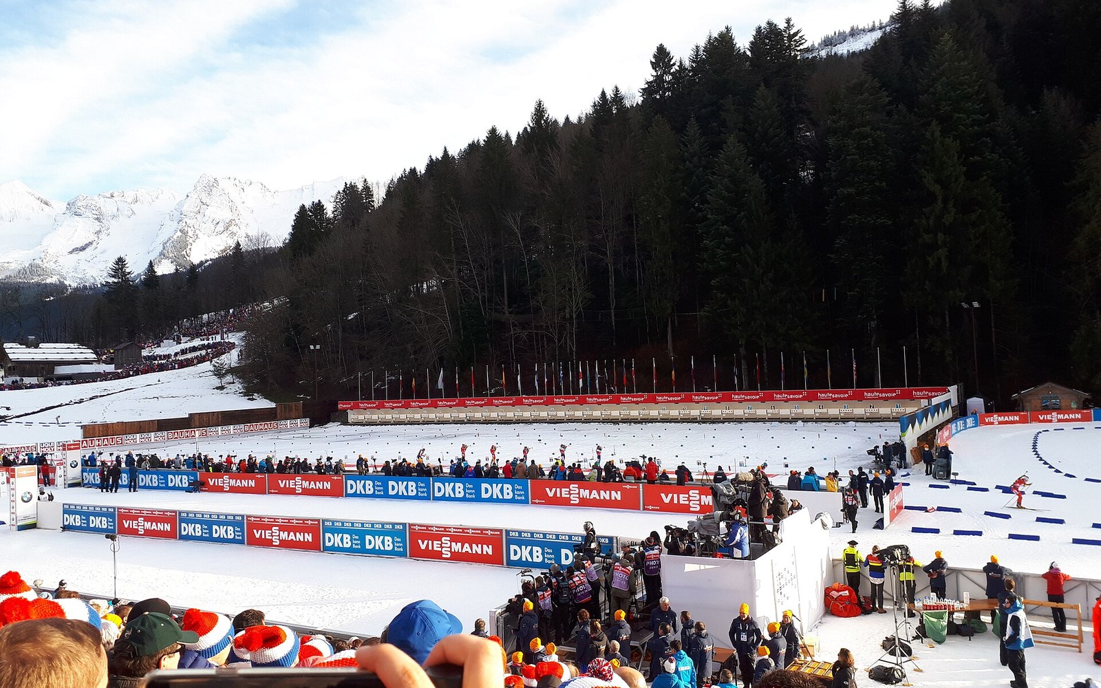 Spectators and officials watch a biathlon shooting range surrounded by snowy pine-covered mountains