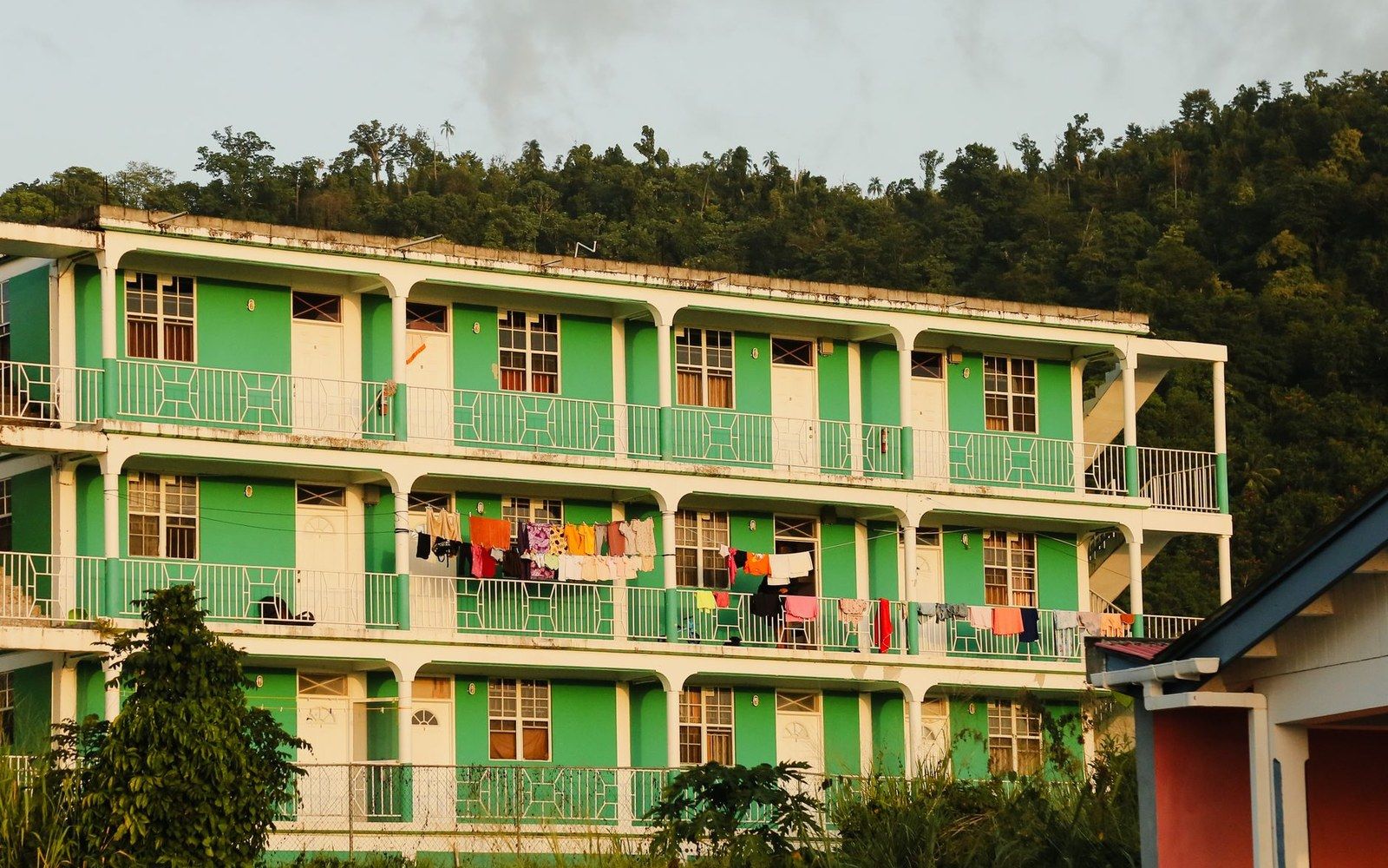 Green three-story balconied apartment building with laundry drying against a forested hillside