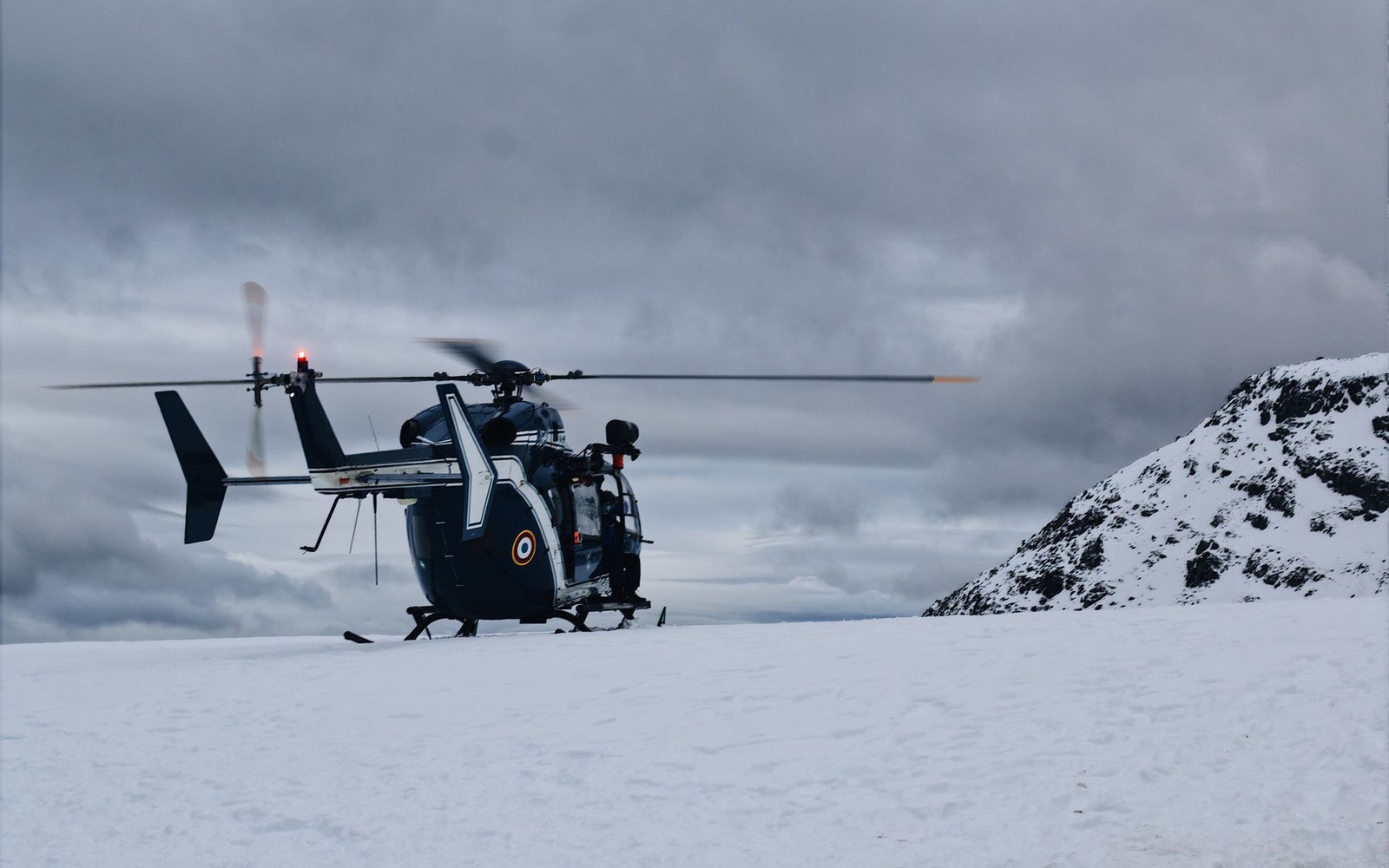 Two PGHM mountain rescue gendarmes in operational gear beside a helicopter on a high-altitude landing pad, the valley far below