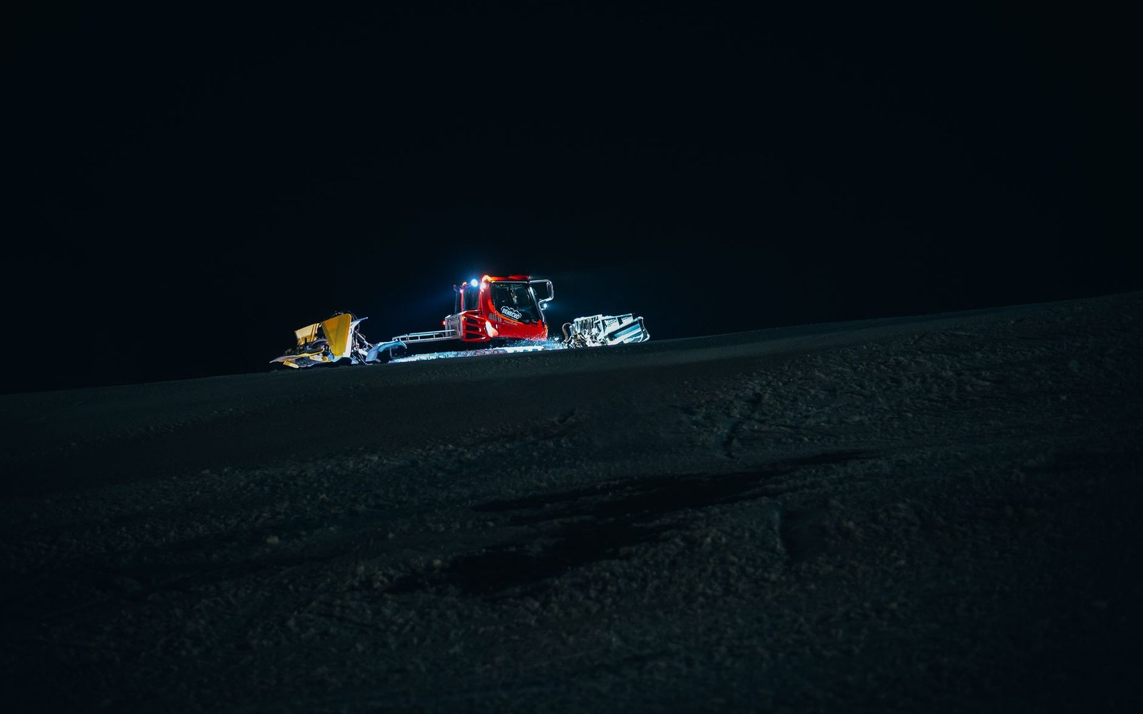 A piste basher with its cab lights on at dusk, working a groomed run with the mountain silhouette behind it, operator visible in the cab